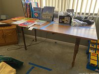 Wooden table with multiple books and boxed items on top, placed near window with vertical blinds.