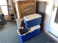 Two stacked blue and white Coleman coolers, medium one on top and large one below. Various other items behind including framed map.