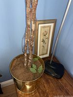 Close-up of faux tree trunk in gold-tone metal pot with soil and leaves, part of lamp base visible, small framed floral picture leaning on wall behind.