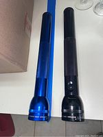 Two large Maglite flashlights, one blue and one black, side by side on white tabletop.