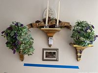 Photo of three gold-colored floating shelves mounted on a light wall. Two hold faux flower arrangements with green leaves and purple flowers, one has two white taper candles and a memorabilia plaque. Below is a framed landscape picture. Shelf design is curved and glossy.
