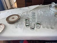 Photo showing variety of glassware including candy dishes, creamer, sugar bowl, several small glasses with some decorated with holly berry design, and a stack of plates with floral design and brown rim.