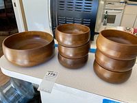 Photo showing all seven items: six medium wooden bowls stacked in two stacks and one large wooden casserole bowl beside them on a kitchen counter.
