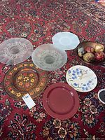 Photo showing six decorative platters of various styles and a metal bowl containing ornaments placed on a patterned rug.