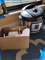 Wide view showing Instant Pot and open cardboard box containing sharpener and cables by the window.
