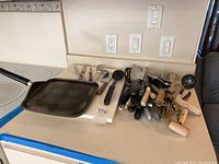 Wide image of countertops with various kitchen utensils, cutters with wooden and metal handles, a metal ladle, forks and cutting board.
