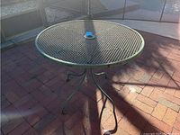 Round metal patio table with lattice top and curved legs on brick patio ground, shows wear.