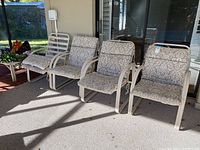 Full view of the four patio chairs lined up against a wall on a beige outdoor carpet. All chairs have matching beige cushions with a floral pattern.