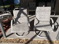 Two beige metal framed swivel outdoor chairs with matching patterned cushions. Chairs are placed outdoors on a rug and exposed to sunlight. Condition appears weathered.