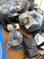 Overview photo of multiple cameras and accessories on table, showing Olympus and Sony cameras, bags, DVDs, and cords.