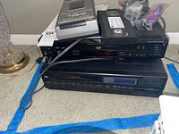 Stacked electronic devices including Sony DVD player and VCR combo, standalone VCR, VCR rewinder, VHS tape, and cables.