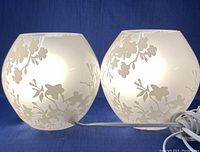 Two frosted glass globe lamps with white cherry blossom floral pattern displayed side by side on a blue background.