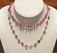 Two beaded necklaces on white display bust. Pink irregular beads with small clear beads necklace and multi-colored orange, purple, and light yellow beads necklace visible.