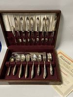 Full view of the wooden chest filled with two sets of silver plated flatware including forks, spoons, knives, and some serving utensils, with a certificate or guarantee card visible.