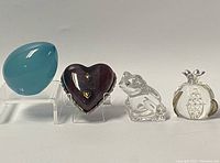 Four vintage glass paperweights displayed in a row including turquoise balloon, burgundy heart, clear glass frog, and pomegranate-shaped items