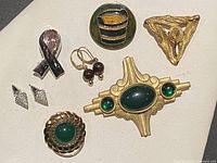Group photo of five vintage costume brooches and two pairs of earrings showing various designs, materials, and colors including green stones, gold-tone metal, purple beads, and silver-tone metal.