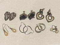 Full lot of six pairs of silver earrings displayed on a light surface including leaf, heart, hoops, black stone studs, and pearl drops.
