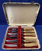 Full view of the open box containing six steak knives with stag antler handles, arranged on red velvet lining.