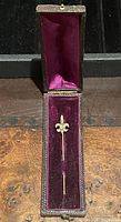 Fleur-de-lis shaped gold stick pin in original velvet-lined display box open vertically showing pin centered inside.