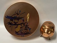 Persian copper tin and copper/brass piggybank shown side by side from above, highlighting the calligraphy and pig's face details.