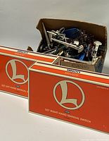 View of two Lionel branded 027 manual switch boxes and a cardboard box with various metal attachments and fixtures for model train set.