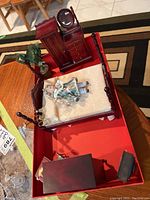 Miniature dollhouse bedroom furniture including bed with cream-colored bedding, dark wood vanity and tall dresser, artificial plant, miniature doll on bed, small chandelier, and additional small chest visible inside red tray.