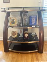 Front view of 4 shelf TV unit with items on shelves for scale only. Unit has black tempered glass shelves, dark wood curved supports, and brushed metal front accents.