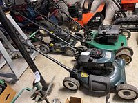 Full side view of the Craftsman lawn mower showing green body, black engine housing and four wheels, placed indoors with other tools and equipment in background.