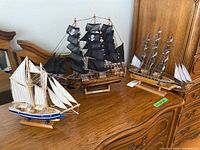 Three model sailing ships shown on a wood cabinet: largest pirate ship Corsaro-Nero with black sails, medium ship with brown striped sails, and smallest blue schooner with cream sails.