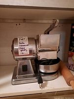 Front view of stainless steel cheese grater on shelf with lever, collecting bowl and power cord visible, wooden rolling pin at side