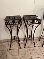 Two matching black metal plant stands with ornate floral and scroll designs. Square lattice tops; one top slightly loose.