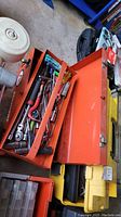 Open red metal toolbox showing various hand tools including wrenches, screwdrivers, socket wrenches, and specialty tools inside compartments and tray.