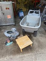 Photo showing plastic wheelbarrow, wooden stool, resin swan planter, and ceramic donkey figure arranged on concrete floor.