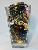 Side view of clear glass jar filled with assorted costume jewelry showing gold-toned bangles, chains, and beaded bracelets