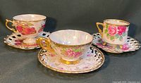 Three hand-painted vintage china teacups and matching pierced edge saucers arranged on dark fabric background.