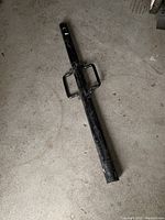 Full view of the black metal post driver with two side handles, lying on concrete floor.