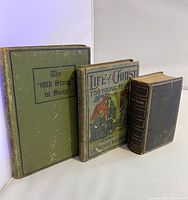 Three antique religious books shown standing upright: The Old Story in Song (green cover), Life of Christ for Young People (illustrated cover), and the black leather-bound Holy Bible with gold edges.