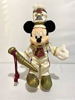 Full front view of Disneyland 50th Anniversary Mickey Mouse plush dressed as parade leader with gold and cream outfit, holding a gold baton with red tassel, wearing tall white hat with 50th emblem.