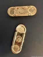 Two oval-shaped metal holders, top view. One holder shows three coins inside with cutouts; the other is solid metal with wear marks.