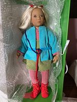 Front view of Annette Himstedt Lottchen doll dressed in blue coat, green dress, red/white striped leggings, red shoes, with blonde hair tied in red ribbons, lying on bubble wrap inside a box.
