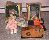 Four Cabbage Patch dolls, two boxed, two unboxed. Two child's western style vests in front. Dolls in varied outfits and styles.
