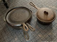Image of three stacked cast iron skillets with one cast iron saucepan with lid next to them showing overall condition and grouping.