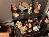 Overall view of a collection of rooster figurines displayed on two-tier wooden furniture. Varied materials and sizes visible.