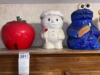 Three cookie jars on wooden shelf: red apple, Pillsbury Doughboy, and Cookie Monster.