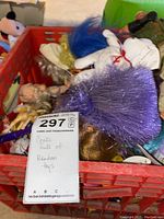 Crate filled with assorted collectible toys including dolls with colorful hair, a white stuffed animal, and an action figure in military gear.