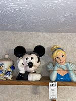 Shelf display of three Disney-themed cookie jars: Donald Duck jar with two small figurines on the left, Mickey Mouse jar in the center, Cinderella jar on the right.