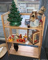 Full front view of the foldable wooden bookshelf with three shelves, holding various ceramic and decorative items including a green ceramic Christmas tree, ceramic dog figure, and other small decorative objects.