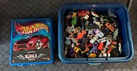 Blue plastic tote filled with many small toy cars and trucks next to a blue carrying case with Hot Wheels graphic lid
