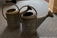 Two galvanized metal watering cans positioned side by side on carpeted floor, showing overall shape and size.