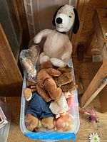 The lot of plush animals and dolls inside a plastic container with various types of dolls and stuffed animals including a large Snoopy plush.